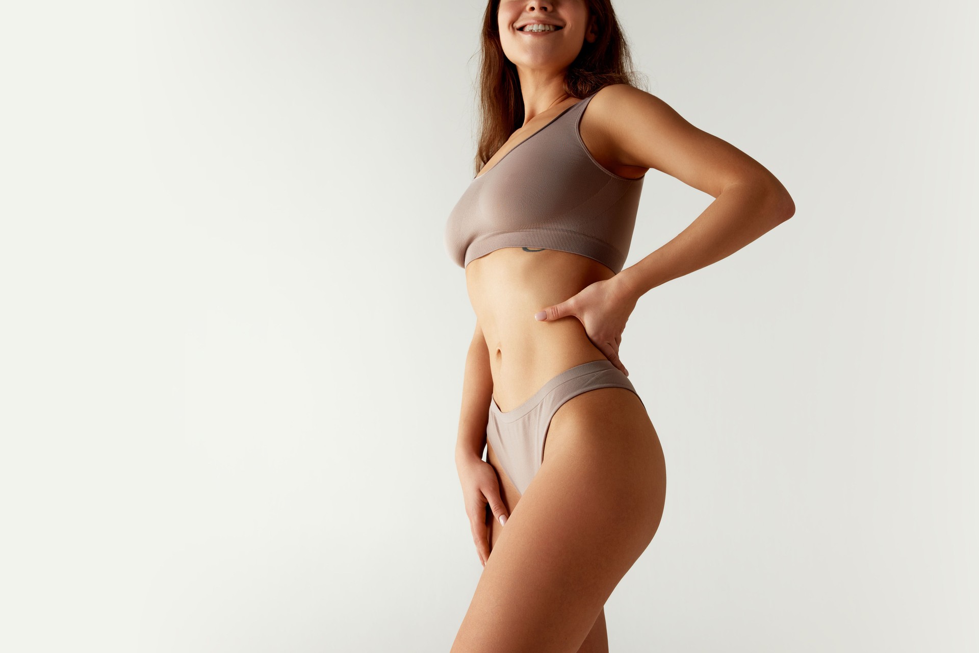 Cropped portrait of woman in sports bra and underwear set, confidently placing hands on her hips against grey studio background.